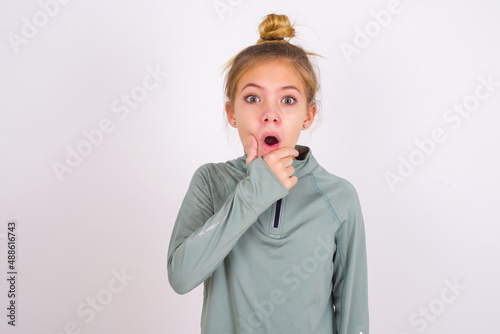 little caucasian kid girl with hair bun wearing technical shirt over white background Looking fascinated with disbelief  surprise and amazed expression with hands on chin