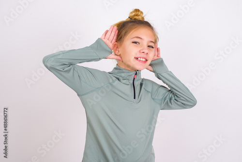 little caucasian kid girl with hair bun wearing technical shirt over white background Trying to hear both hands on ear gesture, curious for gossip. Hearing problem, deaf