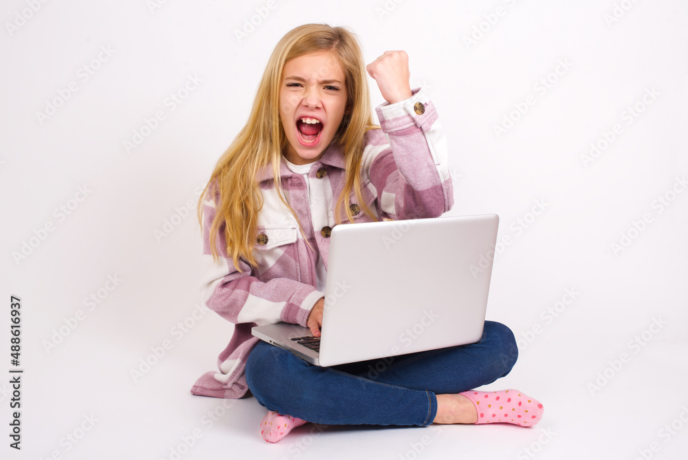 Fierce caucasian teen girl sitting with laptop in lotus position on white background holding fist in front as if is ready for fight or challenge, screaming and having aggressive expression on face. Fierce caucasian teen girl sitting with laptop in lotus position on white background holding fist in front as if is ready for fight or challenge, screaming and having aggressive expression on face.