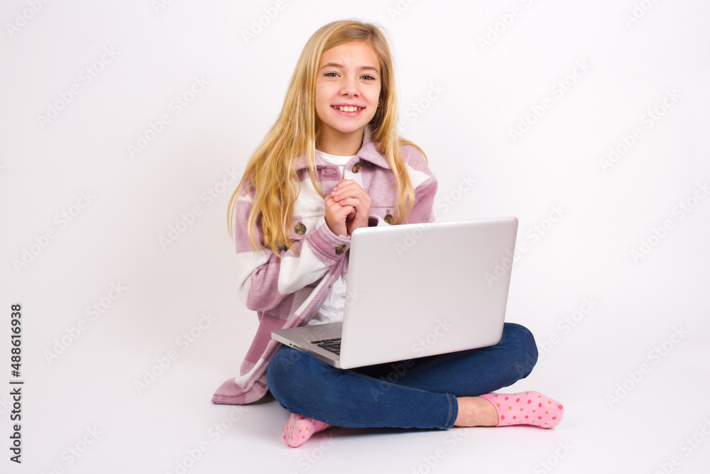Happy caucasian teen girl sitting with laptop in lotus position on white background stands against orange studio wall keeps hands on heart, swears be loyal, expresses gratitude. Honesty concept. Happy caucasian teen girl sitting with laptop in lotus position on white background stands against orange studio wall keeps hands on heart, swears be loyal, expresses gratitude. Honesty concept.