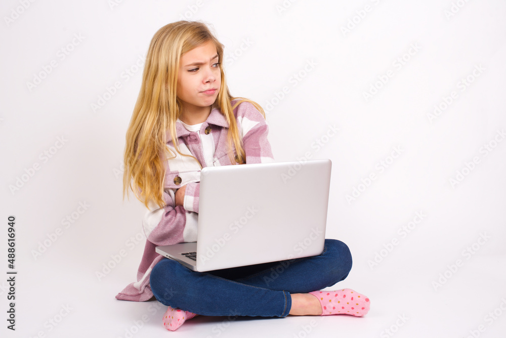Displeased caucasian teen girl sitting with laptop in lotus position on white background with bad attitude, arms crossed looking sideways. Negative human emotion facial expression feelings. Displeased caucasian teen girl sitting with laptop in lotus position on white background with bad attitude, arms crossed looking sideways. Negative human emotion facial expression feelings.