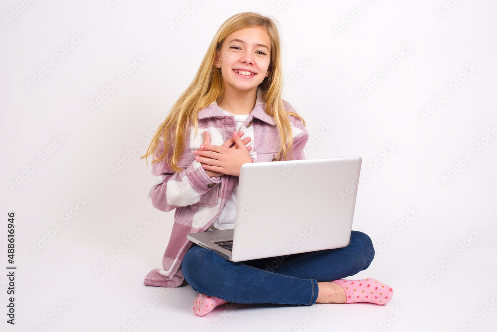 Honest caucasian teen girl sitting with laptop in lotus position on white background keeps hands on chest, touched by compliment or makes promise, looks at camera with great pleasure. Honest caucasian teen girl sitting with laptop in lotus position on white background keeps hands on chest, touched by compliment or makes promise, looks at camera with great pleasure.
