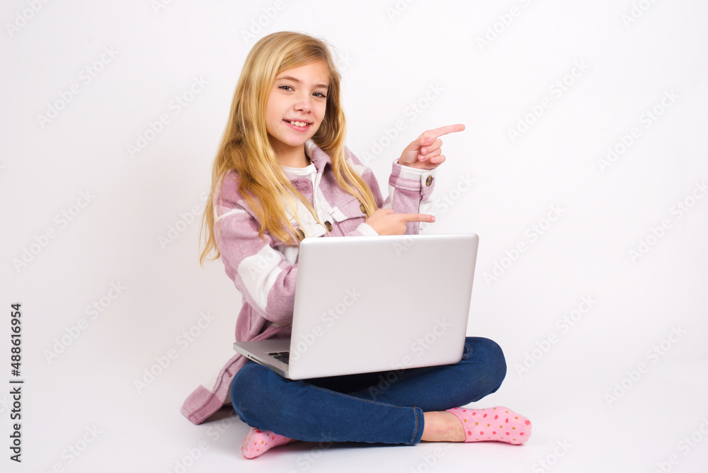 caucasian teen girl sitting with laptop in lotus position on white background points aside with surprised expression with mouth opened, shows something amazing. Advertisement concept. caucasian teen girl sitting with laptop in lotus position on white background points aside with surprised expression with mouth opened, shows something amazing. Advertisement concept.