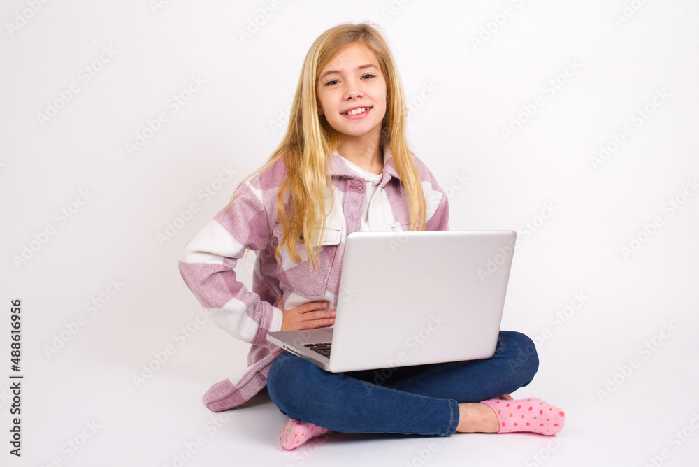 Studio shot of cheerful caucasian teen girl sitting with laptop in lotus position on white background keeps hand on hip, smiles broadly. Studio shot of cheerful caucasian teen girl sitting with laptop in lotus position on white background keeps hand on hip, smiles broadly.