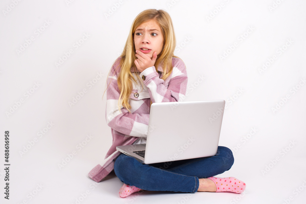 caucasian teen girl sitting with laptop in lotus position on white background covering mouth with hands scared from something or someone bitting nails caucasian teen girl sitting with laptop in lotus position on white background covering mouth with hands scared from something or someone bitting nails