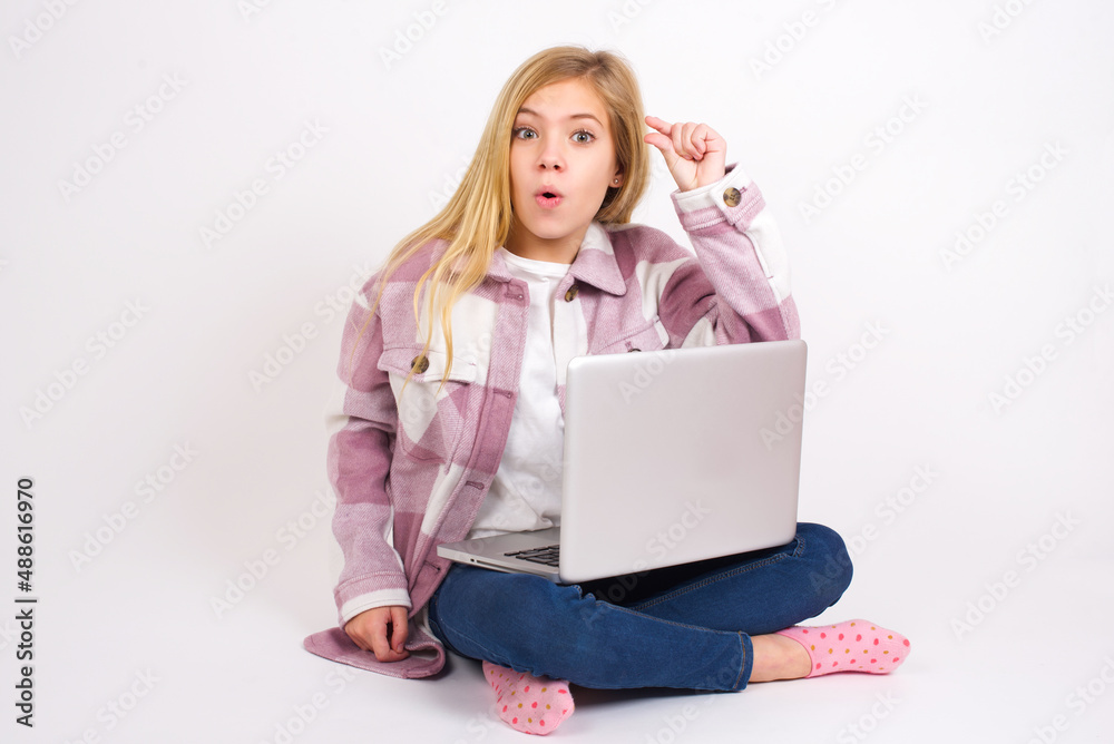 Naklejka premium Shocked caucasian teen girl sitting with laptop in lotus position on white background shows something little with hands, demonstrates size, opens mouth from surprise. Measurement concept.