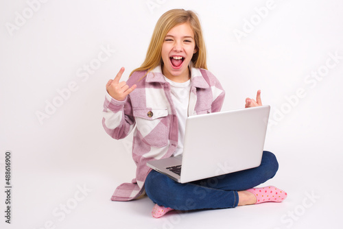 Born to rock this world. Joyful caucasian teen girl sitting with laptop in lotus position on white background screaming out loud and showing with raised arms horns or rock gesture. © Jihan