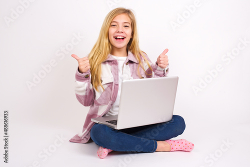 Optimistic caucasian teen girl sitting with laptop in lotus position on white background showing thumbs up with positive emotions. Quality and recommendation concept. © Jihan