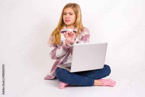 Ugh how disgusting! Displeased caucasian teen girl sitting with laptop in lotus position on white background , has dissatisfied facial expression as sees something abominable. © Jihan
