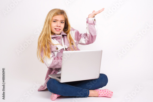 caucasian teen girl sitting with laptop in lotus position on white background pointing aside with both hands showing something strange and saying: I don't know what is this. Advertisement concept. © Jihan