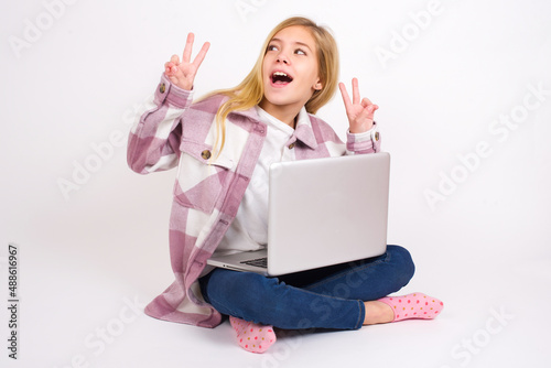 Isolated shot of cheerful caucasian teen girl sitting with laptop in lotus position on white background makes peace or victory sign with both hands, feels cool. © Jihan