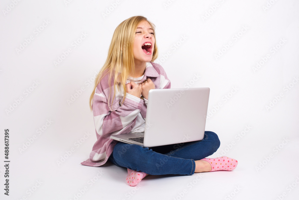 caucasian teen girl sitting with laptop in lotus position on white background excited and glad to achieve victory, clenches fists, screams in excitement with closed eyes,successful person. caucasian teen girl sitting with laptop in lotus position on white background excited and glad to achieve victory, clenches fists, screams in excitement with closed eyes,successful person.