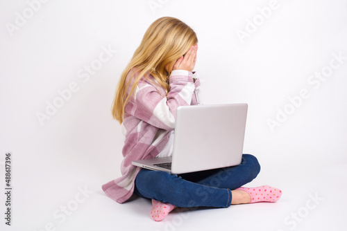 Sad caucasian teen girl sitting with laptop in lotus position on white background covering face with hands and crying. © Jihan