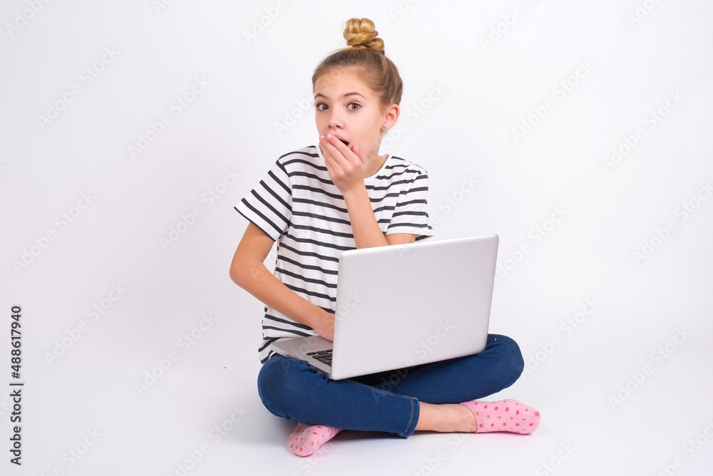 caucasian teen girl sitting with laptop in lotus position on white background covers mouth and looks with wonder at camera, cannot believe unexpected rumors.