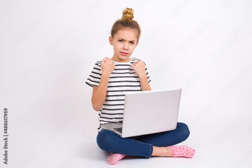 Displeased annoyed caucasian teen girl sitting with laptop in lotus position on white background clenches fists, gestures pissed, ready to revenge, looks with aggression at camera stands full of hate,
