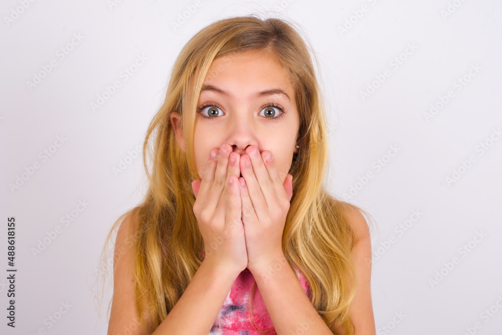 little caucasian kid girl wearing sport clothing over white background keeps hands on mouth, looks with eyes full of disbelief, being puzzled with amount of work little caucasian kid girl wearing sport clothing over white background keeps hands on mouth, looks with eyes full of disbelief, being puzzled with amount of work