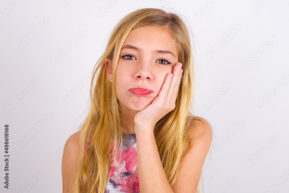 Sad lonely little caucasian kid girl wearing sport clothing over white background touches cheek with hand bites lower lip and gazes with displeasure. Bad emotions Sad lonely little caucasian kid girl wearing sport clothing over white background touches cheek with hand bites lower lip and gazes with displeasure. Bad emotions
