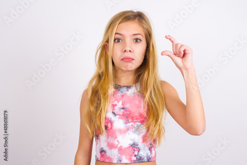 little caucasian kid girl wearing sport clothing over white background purses lip and gestures with hand, shows something very little.