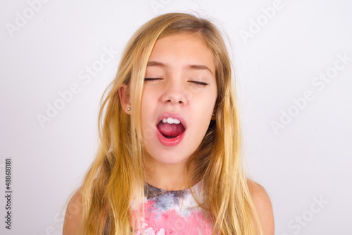 little caucasian kid girl wearing sport clothing over white background yawns with opened mouth stands. Daily morning routine