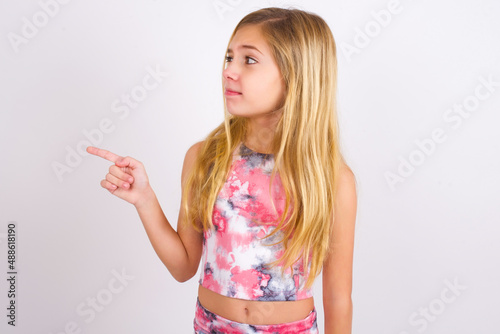 little caucasian kid girl wearing sport clothing over white background points at copy space and advertises something  advices best price.