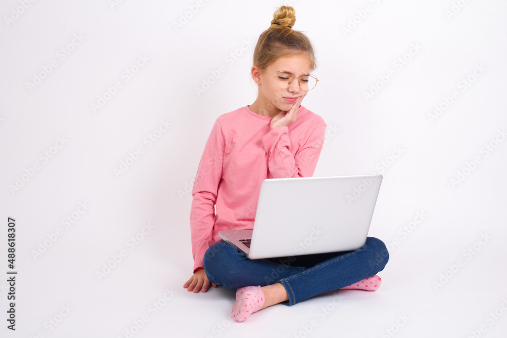 Beautiful caucasian teen girl sitting with laptop in lotus position on white background with toothache Beautiful caucasian teen girl sitting with laptop in lotus position on white background with toothache