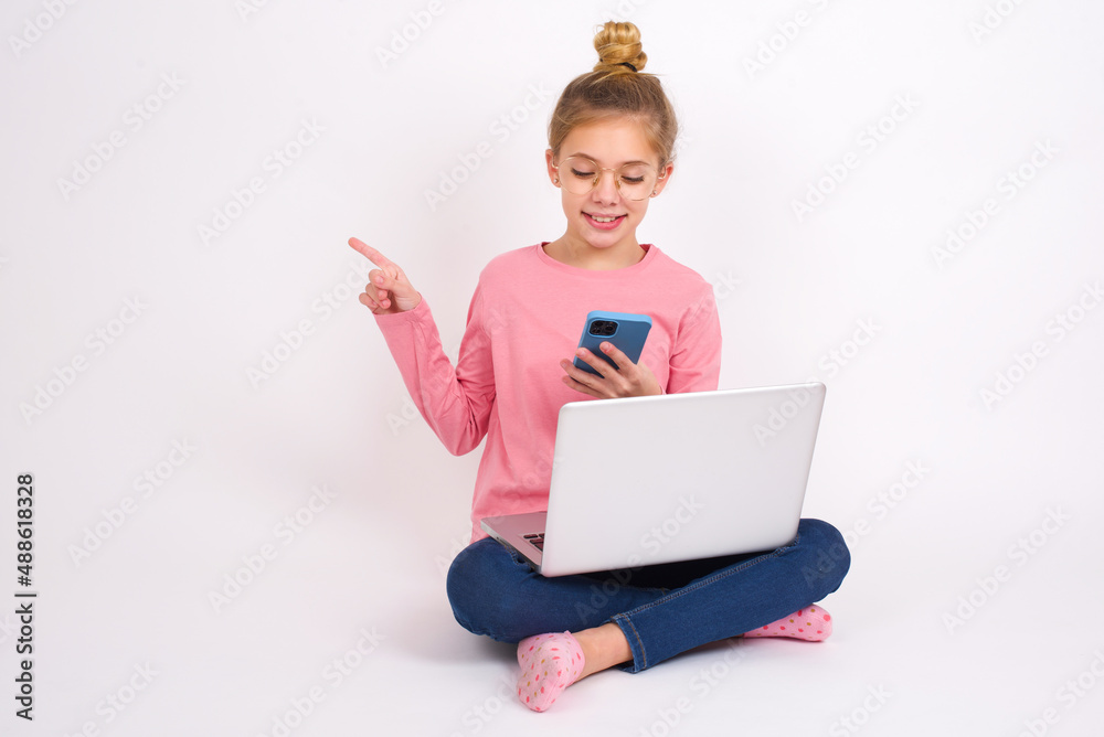 Smiling Beautiful caucasian teen girl sitting with laptop in lotus position on white background pointing finger at blank space holding phone in one hand Smiling Beautiful caucasian teen girl sitting with laptop in lotus position on white background pointing finger at blank space holding phone in one hand