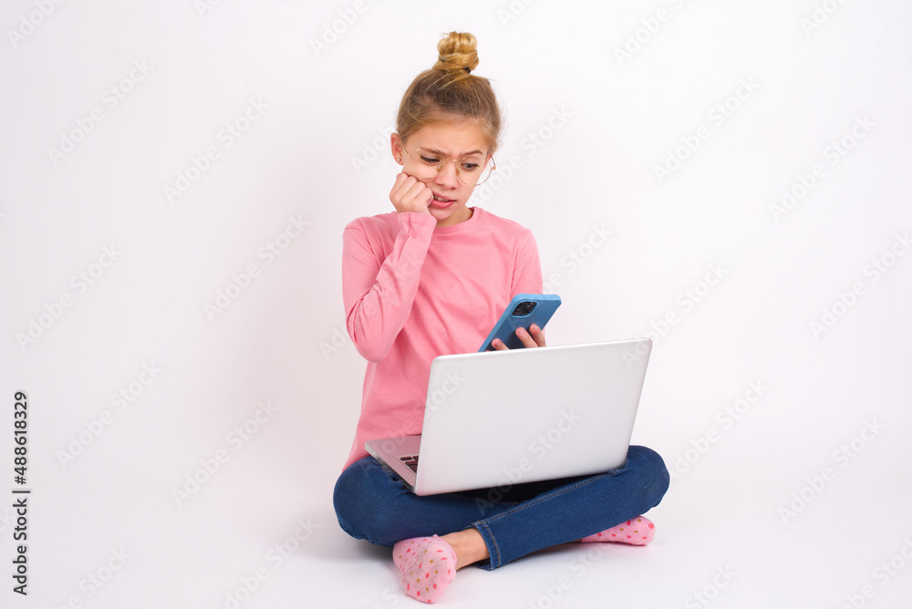Portrait of pretty frightened Beautiful caucasian teen girl sitting with laptop in lotus position on white background chatting biting nails after reading some scary news on her smartphone.