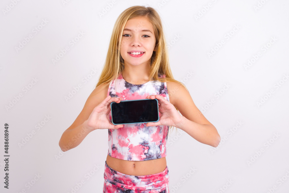 Cheerful cheery content little caucasian kid girl wearing sport clothing over white background holding in hands device hobby smm post blog Cheerful cheery content little caucasian kid girl wearing sport clothing over white background holding in hands device hobby smm post blog