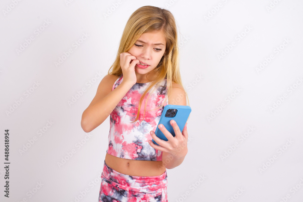 Portrait of pretty frightened little caucasian kid girl wearing sport clothing over white background chatting biting nails after reading some scary news on her smartphone. Portrait of pretty frightened little caucasian kid girl wearing sport clothing over white background chatting biting nails after reading some scary news on her smartphone.