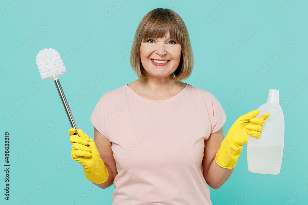 Elderly housewife woman 50s in pink t-shirt gloves hold toilet brush sponge detergent chemical bottle cleanser isolated on plain pastel light blue background. Housekeeping cleaning tidying up concept. Elderly housewife woman 50s in pink t-shirt gloves hold toilet brush sponge detergent chemical bottle cleanser isolated on plain pastel light blue background. Housekeeping cleaning tidying up concept.