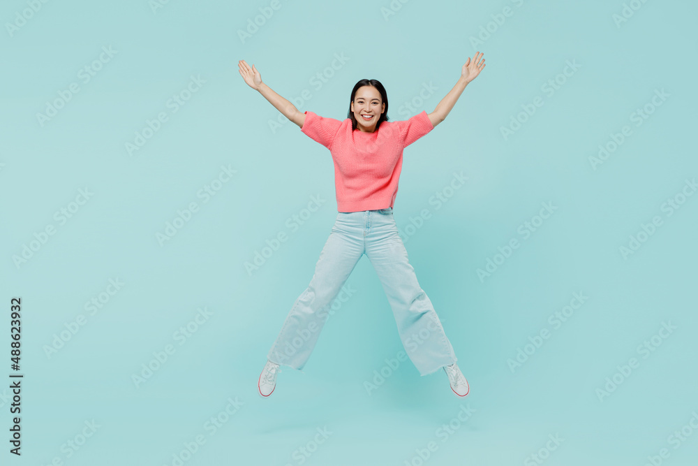 Full body young smiling fun happy woman of Asian ethnicity 20s wearing pink sweater jumpo high with outstretched hands isolated on pastel plain light blue color background. People lifestyle concept.