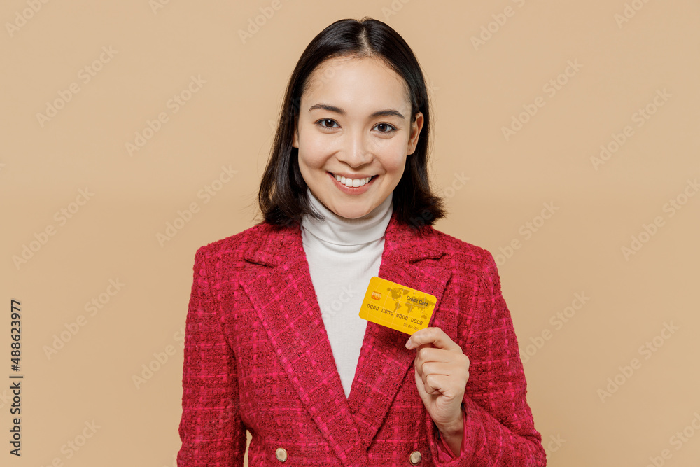 Smiling happy cheerful satisfied woman of Asian ethnicity 20s wear red jacket hold in hand credit bank card isolated on plain pastel beige background studio portrait. People lifestyle fashion concept. Smiling happy cheerful satisfied woman of Asian ethnicity 20s wear red jacket hold in hand credit bank card isolated on plain pastel beige background studio portrait. People lifestyle fashion concept.