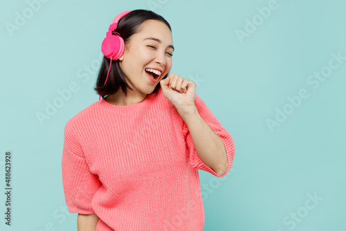 Young smiling happy woman of Asian ethnicity 20s wearing pink sweater headphones listen to music sing song in microphone isolated on pastel plain light blue color background. People lifestyle concept. photo