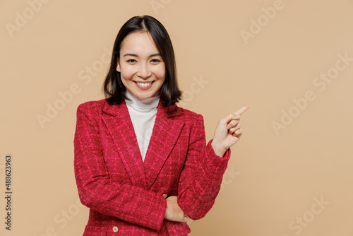 Smiling happy woman of Asian ethnicity 20s in red jacket point index finger aside on workspace area mock up isolated on plain pastel beige background studio portrait. People lifestyle fashion concept. © ViDi Studio