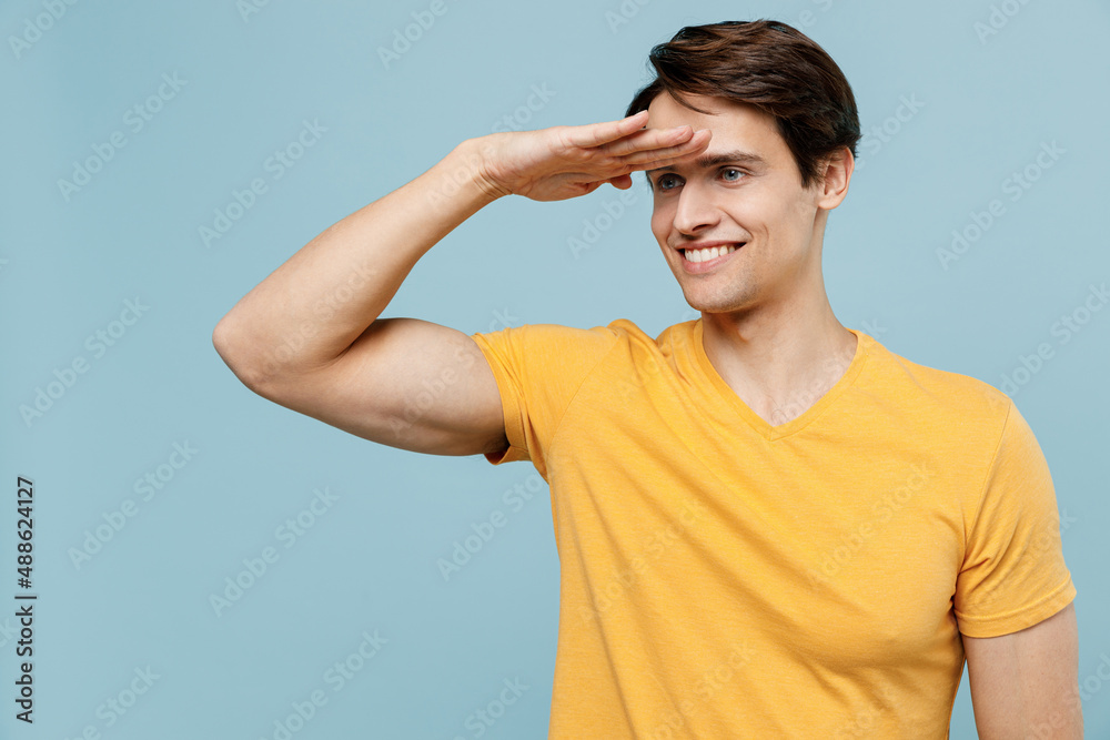 Young smiling happy optimistic cheerful positive man 20s in yellow t-shirt hold hand at forehead look far away distance isolated on plain pastel light blue background studio. People lifestyle concept. Young smiling happy optimistic cheerful positive man 20s in yellow t-shirt hold hand at forehead look far away distance isolated on plain pastel light blue background studio. People lifestyle concept.