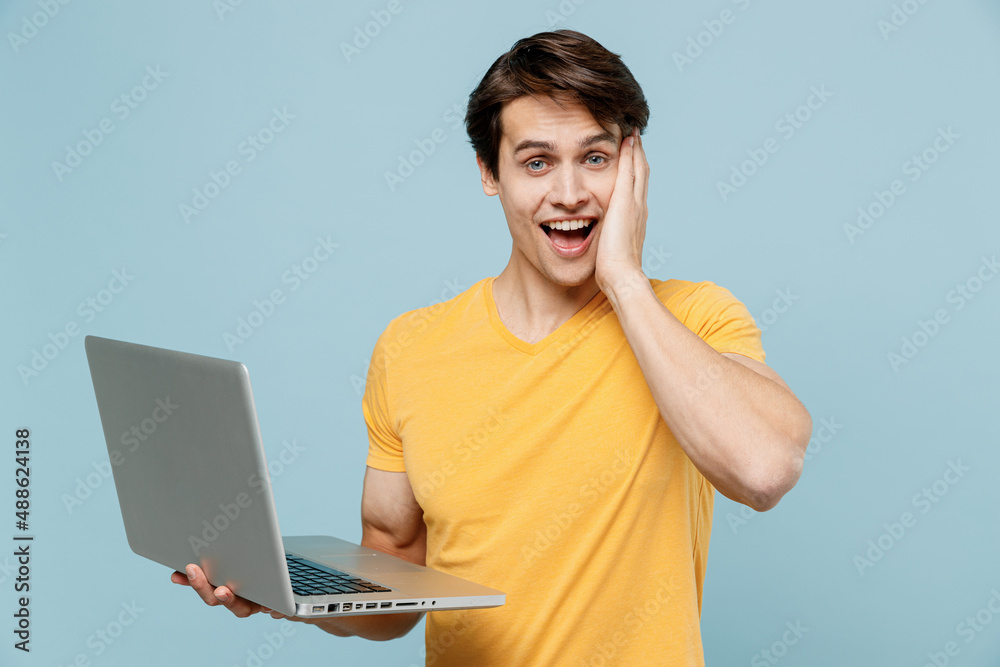 Young amazed happy smiling fun man 20s wearing yellow t-shirt hold use work on laptop pc computer hold face isolated on plain pastel light blue background studio portrait. People lifestyle concept Young amazed happy smiling fun man 20s wearing yellow t-shirt hold use work on laptop pc computer hold face isolated on plain pastel light blue background studio portrait. People lifestyle concept