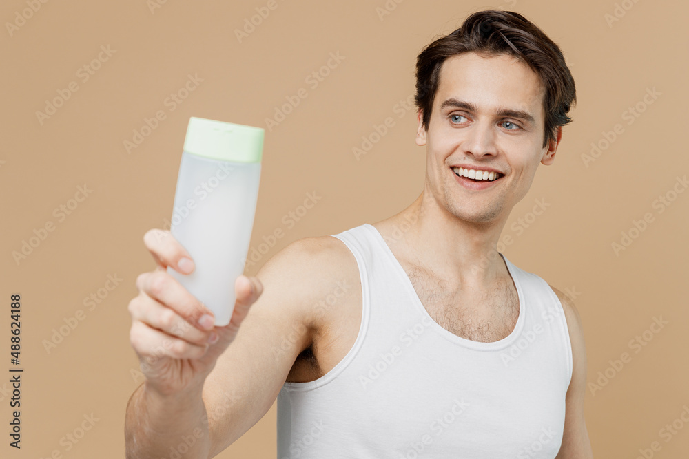 Attractive smiling fun young man 20s perfect skin in undershirt hold in hand close up bottle tonic shampoo isolated on pastel pastel beige background Skin care healthcare cosmetic procedures concept Attractive smiling fun young man 20s perfect skin in undershirt hold in hand close up bottle tonic shampoo isolated on pastel pastel beige background Skin care healthcare cosmetic procedures concept