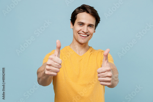 Obraz na plátně Young smiing satisfied cool fun happy cheerful man 20s wear yellow t-shirt show thumb up like gesture validate isolated on plain pastel light blue background studio portrait