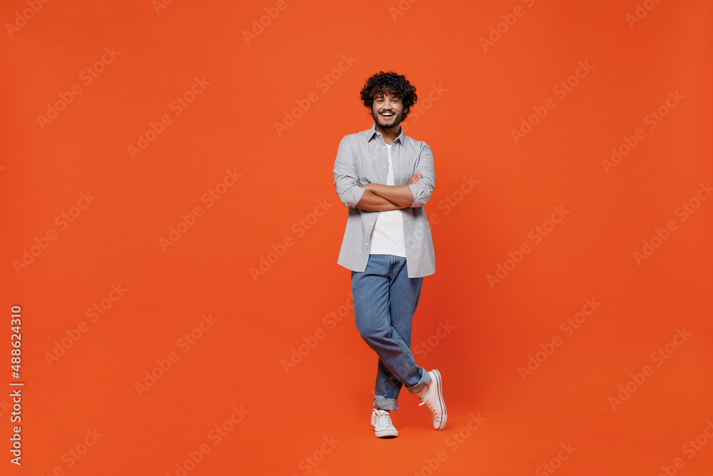 Full size body length confident happy young bearded Indian man 20s years old wears blue shirt hold hands crossed isolated on plain orange background studio portrait. People emotions lifestyle concept.