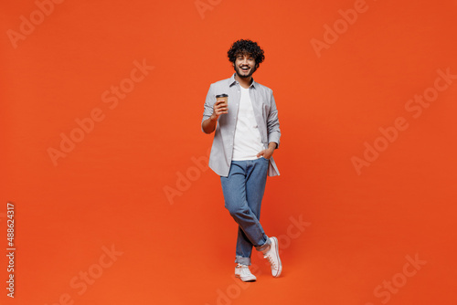 Full size body length fascinating young bearded Indian man 20s years old wears blue shirt hold takeaway delivery craft paper brown cup coffee to go isolated on plain orange background studio portrait.