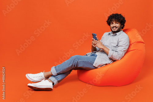 Full size body length young bearded Indian man 20s years old wears blue shirt sit in bag chair hold in hand use mobile cell phone chatting send sms isolated on plain orange background studio portrait.