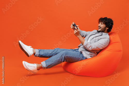 Full size body length vivid young bearded Indian man 20s years old wear blue shirt sit in bag chair hold in hand play pc game with joystick console isolated on plain orange background studio portrait photo