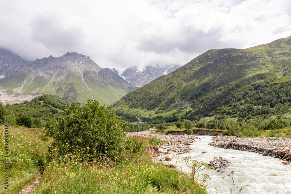 Patara Enguri River flowing down the a valley in the Greater Caucasus Mountain Range in Georgia, Svaneti Region, Ushguli. The Shkhara Mountain is covered by clouds. Snow-capped mountains.Glacier Patara Enguri River flowing down the a valley in the Greater Caucasus Mountain Range in Georgia, Svaneti Region, Ushguli. The Shkhara Mountain is covered by clouds. Snow-capped mountains.Glacier