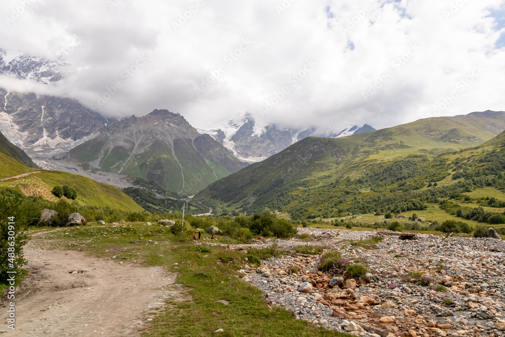 A hiking path leading to the Shkhara Glacier in the Greater Caucasus Mountain Range in Georgia, Svaneti Region, Ushguli. Snow-capped mountains in the back. Wanderlust. Wilderness. Overcast and clouds