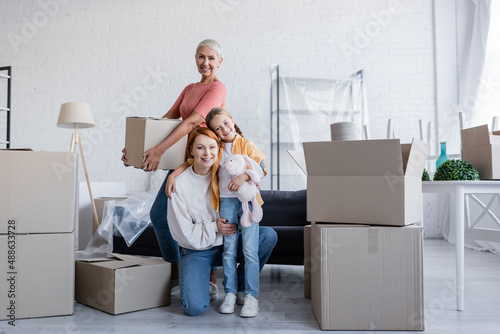 joyful same sex couple with adopted daughter looking at camera near carton packages in new home photo