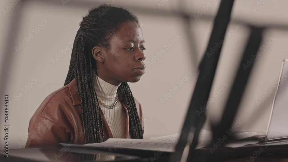 Waist up slowmo of young African-American woman learning to play piano having online video lesson on laptop studying musical notes together with teacher