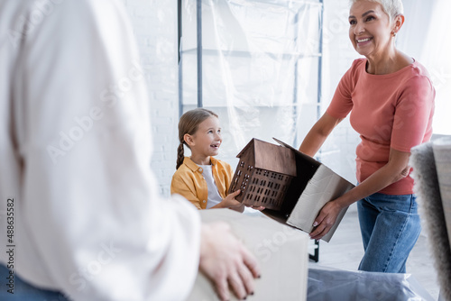 cheerful child holding house model near same sex parents with carton boxes