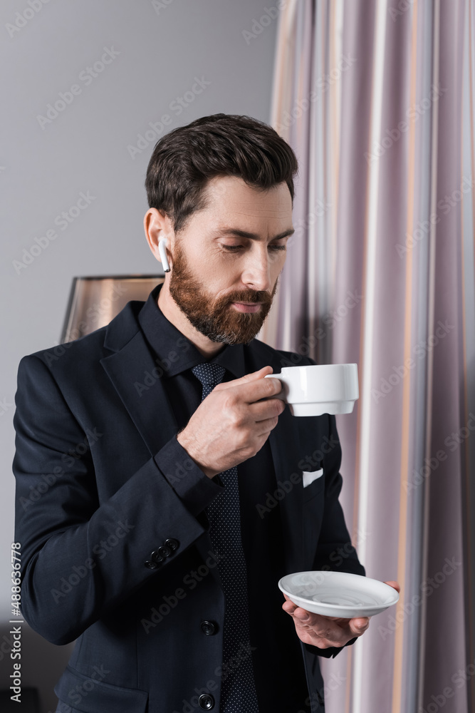 bearded businessman in earphone holding cup and saucer in hotel room bearded businessman in earphone holding cup and saucer in hotel room