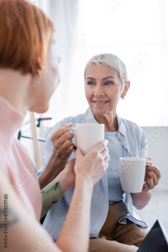 blurred woman taking cup of tea from smiling lesbian girlfriend