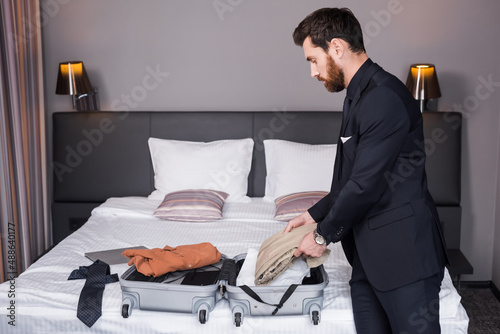 Fotografia side view of bearded man in suit unpacking suitcase in hotel room